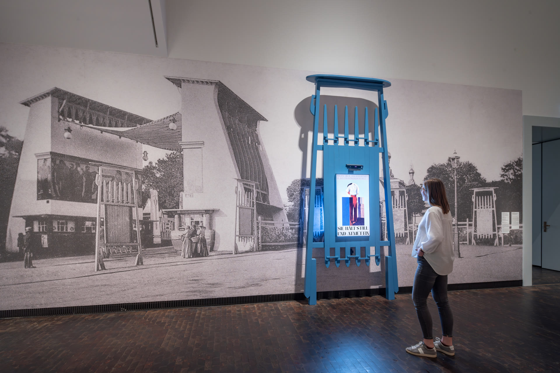 Visitor standing in front of the blue kiosk as it generates their poster, against the Mathildenhöhe mural backdrop.