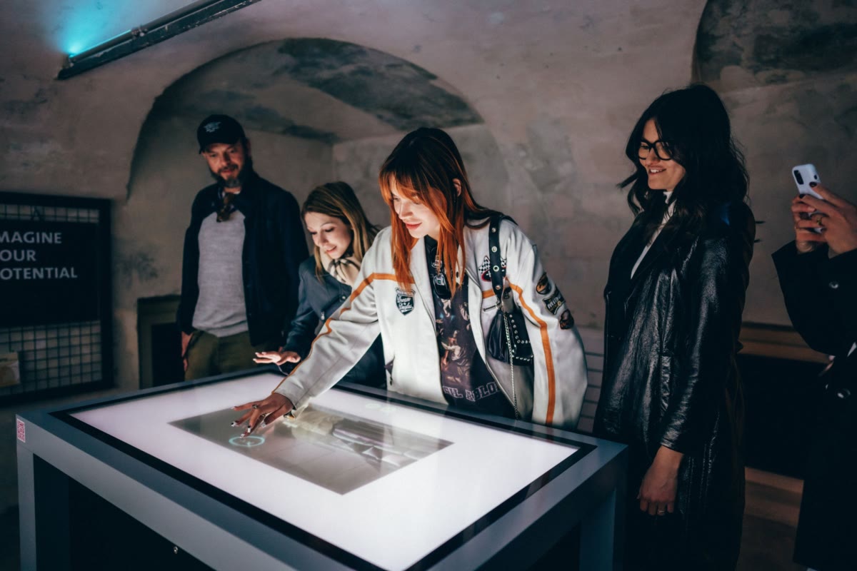 A small crowd gathered around the touchscreen table in a vaulted stone room, with an 'Imagine Your Potential' wall sign in the background.