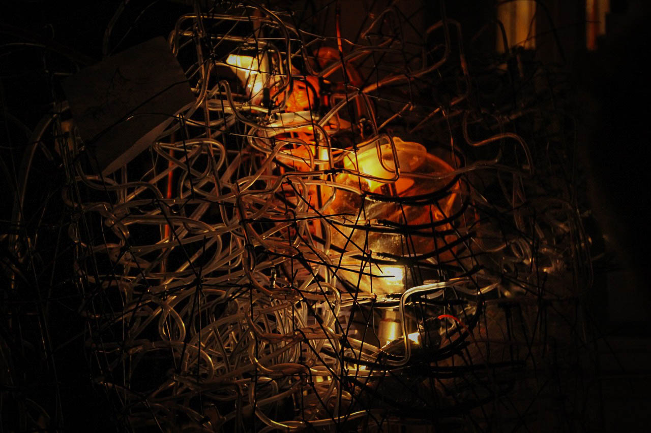 Amber light glowing at the sculpture's core, seen through the dense wire and tubing frame.