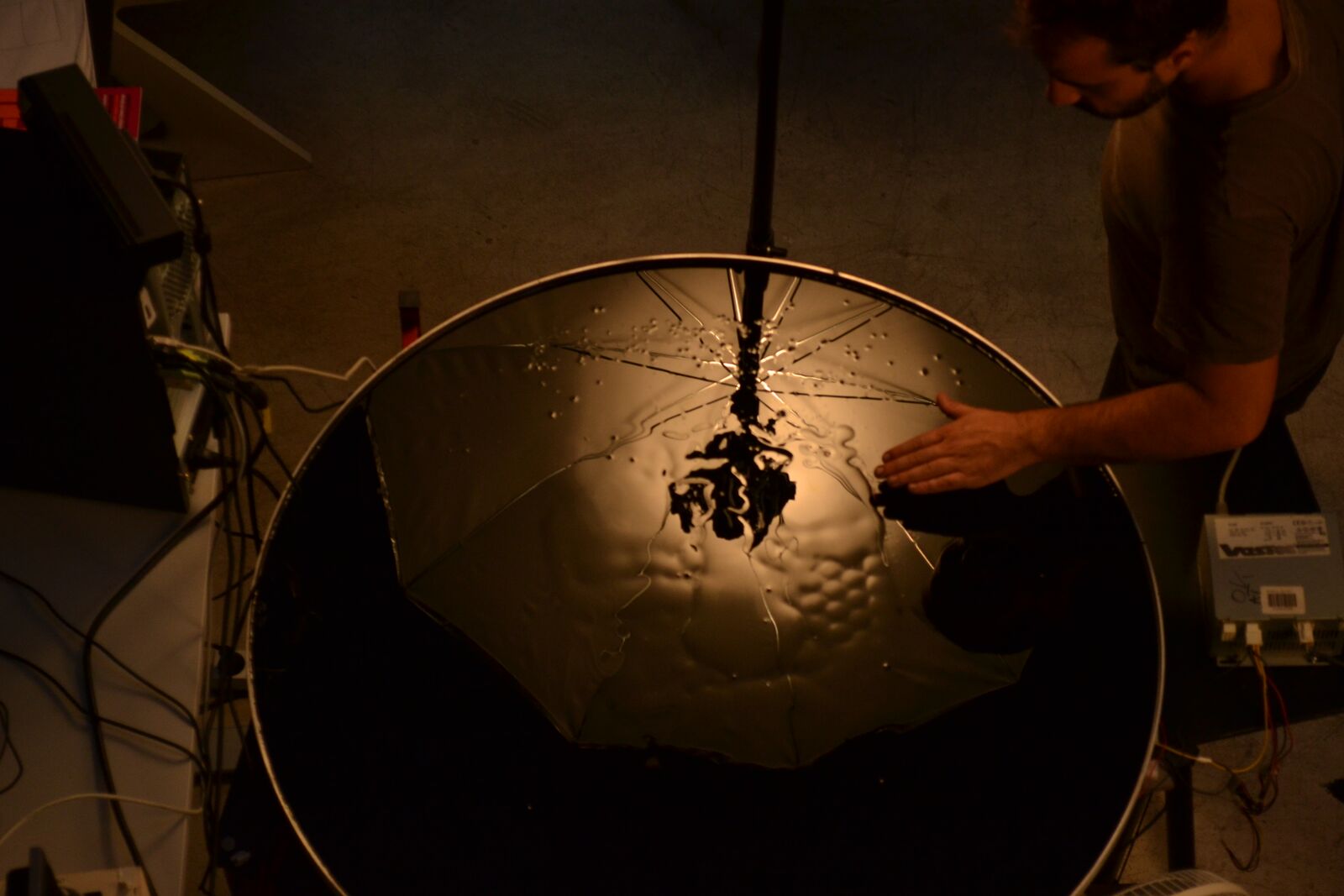Overhead view of the ferrofluid tank, a dark filament of fluid rising from the surface as a hand reaches toward it.